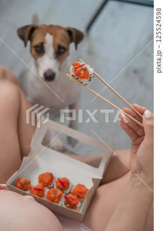 A pregnant woman sits on the sofa and eats rolls. Jack Russell Terrier dog sits on the floor and begs for food from its owner. A pregnant woman sits on the sofa and eats rolls. Jack Russell Terrier dog sits on the floor and begs for food from its owner. 125524598