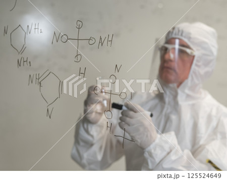 An elderly Caucasian male scientist in a protective suit holds a test tube and writes a formula on the glass.  125524649