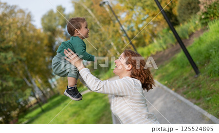 Caucasian woman holds her one-year-old son in her arms while walking in the park in autumn. Caucasian woman holds her one-year-old son in her arms while walking in the park in autumn. 125524895