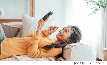 Happy young asian woman relax on comfortable couch at home texting messaging on smartphone 125527283