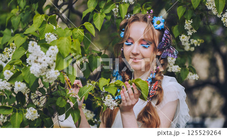 Woman wearing vibrant makeup, butterfly hair clips, inhaling fragrant white lilacs amid blossoming garden spring scenery 125529264