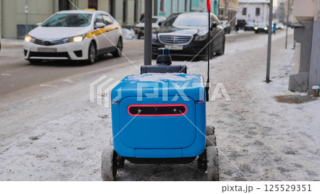 Autonomous delivery robot moving on a snowy sidewalk, navigating urban environment in Moscow. 125529351