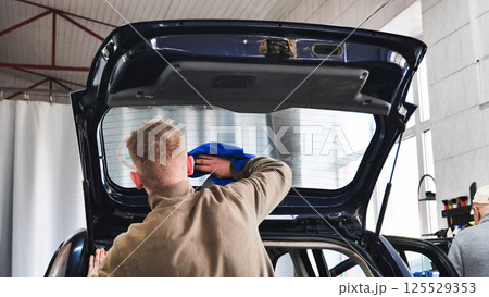 Car wash employee cleaning rear window of vehicle with open trunk 125529353