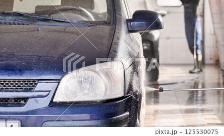 Car wash worker cleaning dirty blue car with high pressure water jet 125530075