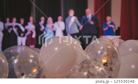 White, transparent balloons filled with gold, silver confetti highlighting blurred graduating students and teachers standing on background stage 125530148