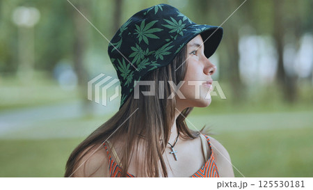 Teenage girl sporting cannabis patterned bucket hat, relaxing outdoors during sunny summer afternoon Teenage girl sporting cannabis patterned bucket hat, relaxing outdoors during sunny summer afternoon 125530181