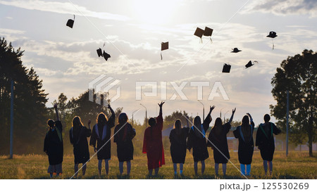 Happy students in graduation gowns toss their caps into the air against a beautiful sunset background Happy students in graduation gowns toss their caps into the air against a beautiful sunset background 125530269