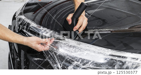 Car wrapping specialist putting vinyl foil or film on car. Protective film on the car. Applying a protective film to the car with tools. Car detailing. Transparent film. Car paint protection. Trimming 125530377