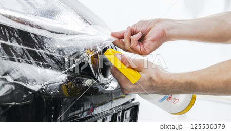 Car wrapping specialist putting vinyl foil or film on car. Protective film on the car. Applying a protective film to the car with tools. Car detailing. Transparent film. Car paint protection. Trimming 125530379