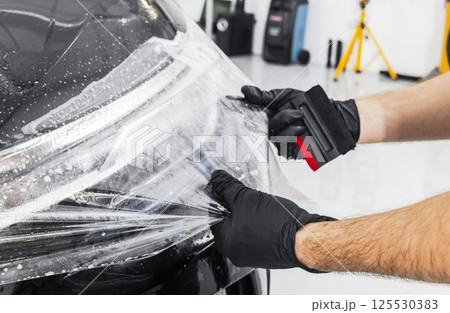 Car wrapping specialist putting vinyl foil or film on car. Protective film on the car. Applying a protective film to the car with tools. Car detailing. Transparent film. Car paint protection. Trimming 125530383