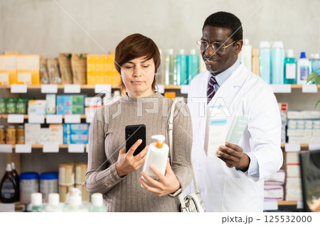 Woman in pharmacy scans QR code on body cream. African man pharmacist in background 125532000