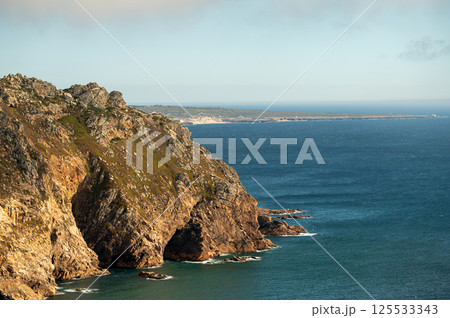 The sun casts a warm glow on rugged cliffs above tranquil waters with a misty backdrop. The sun casts a warm glow on rugged cliffs above tranquil waters with a misty backdrop. 125533343