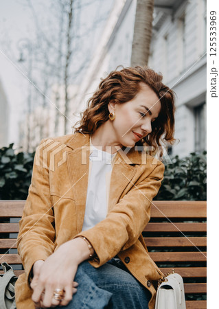 A young woman with curly red hair sits quietly on a bench looking down with a calm expression. Quiet contemplation, thoughtful mood, inner calm, soft elegance... 125533969