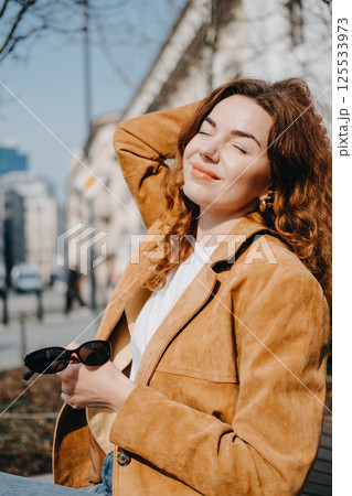 Young woman with auburn curly hair enjoying sunlight on a city street bench. Urban mindfulness, mental wellness, self-care, emotional well-being... 125533973