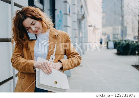 A young woman multitasks with her phone while opening her bag on a city sidewalk. Mobile work culture, communication on the move, hybrid professional life, flexible workspace. 125533975