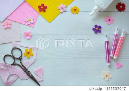 Flat lay composition of DIY greeting card crafting materials, including colorful felt flowers, scissors, glue, arranged on a white wooden background, showcasing the process of creating a personalized 125537947