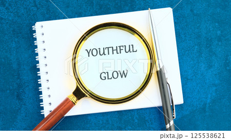A magnifying glass highlights the phrase youthful glow on a notepad, symbolizing the pursuit of vitality and beauty through self-care. A pen rests nearby, ready for notes A magnifying glass highlights the phrase youthful glow on a notepad, symbolizing the pursuit of vitality and beauty through self-care. A pen rests nearby, ready for notes 125538621