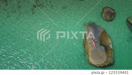 A person lays on a rock in the middle of calm turquoise water, surrounded by scattered stones. The sunlight illuminates the scene, creating a peaceful atmosphere. 125539481