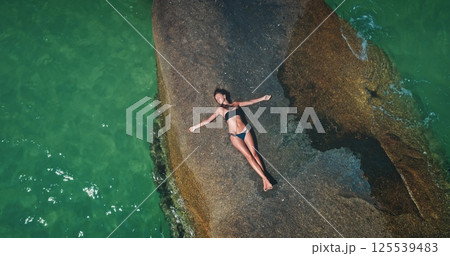 A woman lies on a flat rock surface surrounded by clear turquoise water. Sunlight enhances the vibrant colors as she enjoys a peaceful moment in a tropical beach setting. 125539483