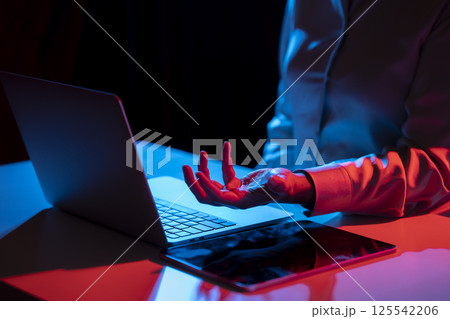 Businesswoman with laptop computer and tablet on the table, holding or showing something in her hand. Mockup and copy space. Business and technology concept. Illuminated with blue and red light 125542206