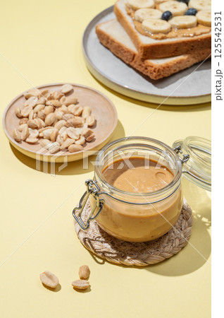 Peanut butter in glass jar on yellow background with salted nuts, toast, banana and berries. Peanut butter in glass jar on yellow background with salted nuts, toast, banana and berries. 125542531