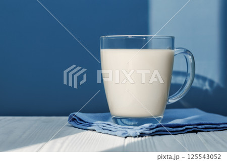 Sunlight illuminating glass milk resting on blue napkin on white wooden table with blue background, creating cozy scene 125543052