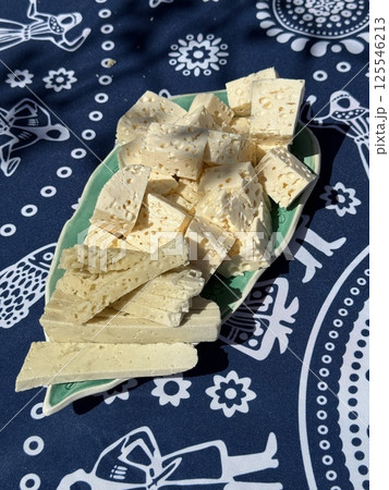 Cubes and strips of porous cheese are neatly arranged on a green-edged plate, placed over a decorative blue cloth with folkloric white patterns. 125546213