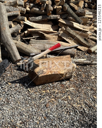A splitting maul embedded in a log, surrounded by chopped and unchopped firewood on gravel under bright sunlight. 125546215