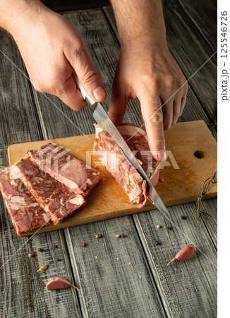 A person slices fresh meat on a rustic wooden cutting board. Surrounding the meat are garlic cloves and fresh herbs, creating a cozy culinary atmosphere A person slices fresh meat on a rustic wooden cutting board. Surrounding the meat are garlic cloves and fresh herbs, creating a cozy culinary atmosphere 125546726