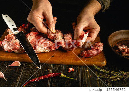 A person is skillfully cutting and preparing fresh meat on a wooden board, surrounded by herbs, garlic, and spices. The kitchen has a warm, inviting atmosphere A person is skillfully cutting and preparing fresh meat on a wooden board, surrounded by herbs, garlic, and spices. The kitchen has a warm, inviting atmosphere 125546732