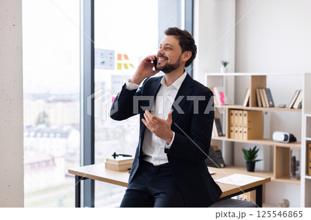 Confident Caucasian male professional in his 30s seated in modern office interior gesturing while conversing on phone, conveying positivity, technology use, communication, work discussion 125546865