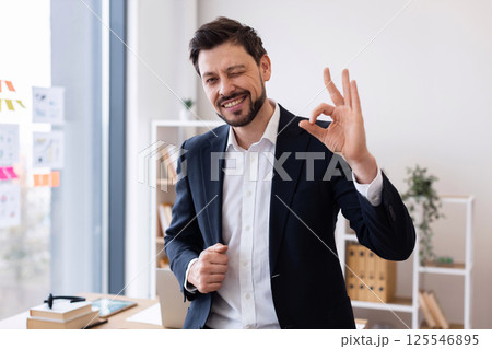 Confident young adult male in formal suit showing okay sign, expressing positivity, working in office environment with window and shelves in background portraying success, engagement, and assurance 125546895