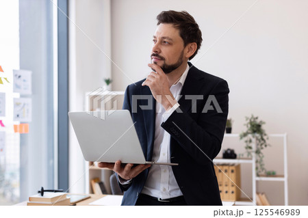 Man in early 30s wearing suit in office, holding laptop, expressing thoughtful pose, confidence. Man in early 30s wearing suit in office, holding laptop, expressing thoughtful pose, confidence. 125546989