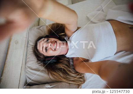 Woman in lingerie listening to music in headphones and smiling in bedroom 125547730