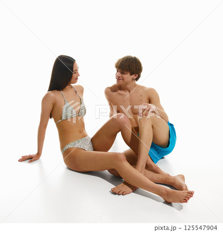 Young woman in swimsuit sitting on floor, talking to muscular shirtless man in swimming trunks against white studio background. Young woman in swimsuit sitting on floor, talking to muscular shirtless man in swimming trunks against white studio background. 125547904
