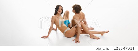 Handsome young man and beautiful woman, couple in swimsuits posing on floor against white studio background. 125547911