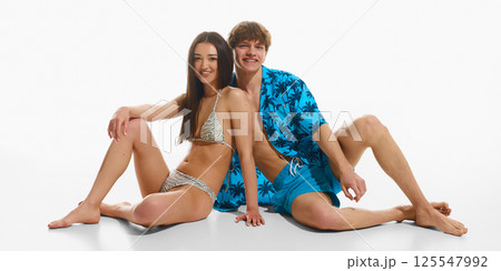 Happy couple sitting barefoot laughing together, man wearing tropical beach wear and woman in elegant swimsuit, posing against white studio background. 125547992