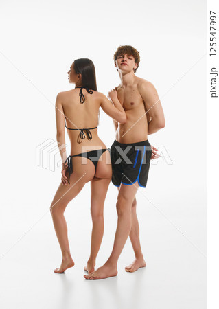 Handsome young man in black swimming trunks posing with elegant young slim woman in black swimsuit, posing against white studio background Handsome young man in black swimming trunks posing with elegant young slim woman in black swimsuit, posing against white studio background 125547997