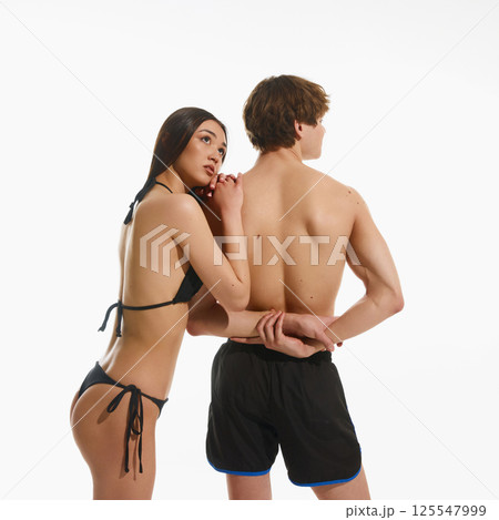 Elegant young woman with fit, tanned body in black swimwear leaning on male muscular back standing in swimming trunks against white background 125547999