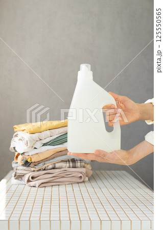 Mockup, woman's hand holding conditioner or laundry gel against a stack of clean clothes.  125550565