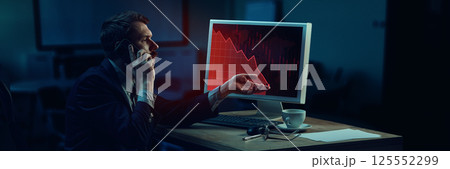 Businessman sitting in modern dark office, arguing on phone, looking at screen showing dramatic stock market crash 125552299