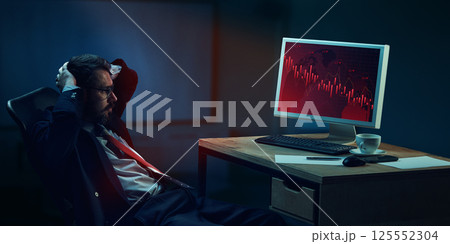 Stressed businessman sitting at desk in modern office late night, with hands behind head, looking at screen showing falling economic graphs 125552304