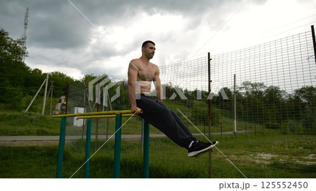 Strong man trains abdominal muscles raising legs on parallel bars at sports ground. Young shirtless sportsman exercising at nature. Sporty athlete working out at nature. Concept of sportive lifestyle Strong man trains abdominal muscles raising legs on parallel bars at sports ground. Young shirtless sportsman exercising at nature. Sporty athlete working out at nature. Concept of sportive lifestyle 125552450
