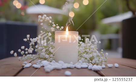 close-up of a white candle with a flame burning brightly on a wooden surface. The candle is placed in the center of the image, surrounded by small white flowers and pebbles	 125555237
