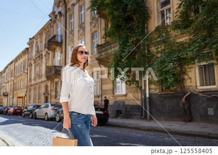 Young woman walking down a sunlit street carrying a shopping bag on a breezy day in a charming neighborhood 125558405