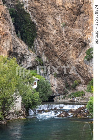 Buna river near the Blagaj Tekke monastery, Bosnia and Herzegovina, Europe. Idyllic scene of a small house nestled at the base of a towering cliff face alongside a rushing river with small waterfalls. 125558974