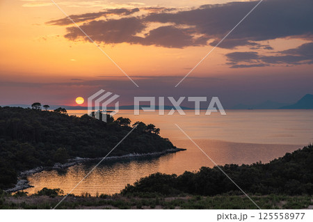 The sea coast in Peljesac peninsula in southern Dalmatia in Croatia, Europe. Sunset over a tranquil bay, with a treelined coast silhouetted against the vibrant sky and reflective water. 125558977