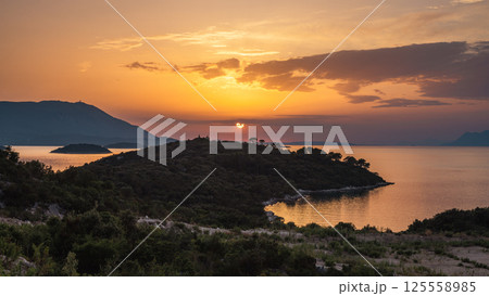 The sea coast in Peljesac peninsula in southern Dalmatia in Croatia, Europe. Golden sunset over a wooded coastline with tranquil waters and distant mountains painted in soft twilight hues. The sea coast in Peljesac peninsula in southern Dalmatia in Croatia, Europe. Golden sunset over a wooded coastline with tranquil waters and distant mountains painted in soft twilight hues. 125558985
