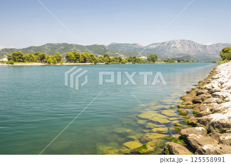 Neretva river near the mouth of river into Adriatic Sea, Croatia, Europe. Scenic waterscape featuring a tranquil river with a rocky shoreline, lush greenery, and distant mountains under a clear sky. 125558991