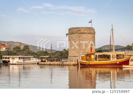 Mali Ston, village in Peljesac peninsula in Croatia, Europe. Picturesque Croatian harbor scene with boats moored near an old stone tower, reflecting in the calm water, under a soft blue sky. 125558997